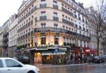 Latin Quarter A bustling Paris street corner in the Latin Quarter features a classic cream-colored building with ornate balconies. At ground level, the lively café “Paris” boasts green awnings, outdoor seating, and people dining as cars and pedestrians navigate the wet street.