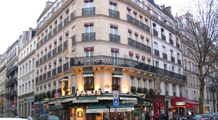 Latin Quarter A bustling Paris street corner in the Latin Quarter features a classic cream-colored building with ornate balconies. At ground level, the lively café “Paris” boasts green awnings, outdoor seating, and people dining as cars and pedestrians navigate the wet street.