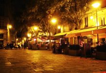 Le Marais A warmly lit outdoor café on a cobblestone street at night. Trees line the street and yellow streetlights illuminate the area. Several people sit at tables under orange awnings. Buildings with lit windows form the backdrop. The atmosphere feels calm and inviting.