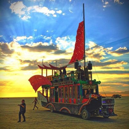 A colorful, ornate vehicle shaped like a Chinese junk boat with red sails stands on a flat desert under a dramatic sunset sky at BurningMan. People are on and around the vehicle as sun rays break through, creating photos impossibles moments.