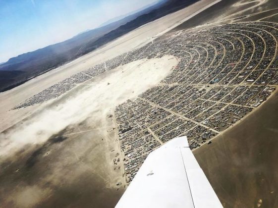 Aerial view of a large desert encampment at BurningMan, arranged in a semi-circular pattern with rows of tents and vehicles. The arid, sandy landscape stretches to distant mountains. Part of an airplane wing is visible in these unique Burning Man photos.