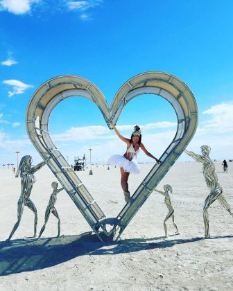 At Burning Man 2024, a woman in a white tutu and headdress balances within a large heart-shaped metal sculpture featuring two adults and two children holding hands. This scene, like many photos impossibles, unfolds on the vast desert under a bright blue sky.