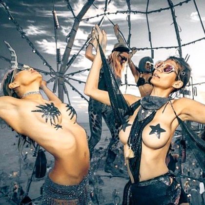 Several women in creative, revealing costumes with body paint and glitter pose expressively at an outdoor BurningMan festival. A large metal structure towers beneath a dramatic cloudy sky, capturing the magic of photos impossibles among fellow festival-goers.