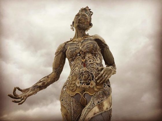 A tall, ornate metallic sculpture of a woman stands against a cloudy sky at BurningMan. Intricately detailed with swirling patterns, gears, and filigree, it blends fantasy and steampunk—a striking figure often seen in unforgettable BurningMan photos.