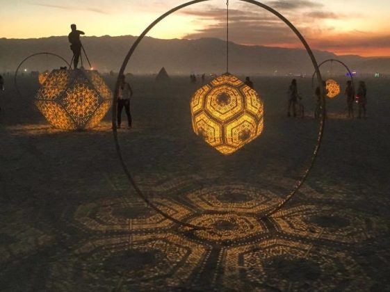 Large geometric lanterns with intricate patterns hang inside hoops, glowing and casting detailed shadows on the sand at dusk. Silhouettes interact with the lanterns, creating magical BurningMan photos against distant mountains and a colorful sky.