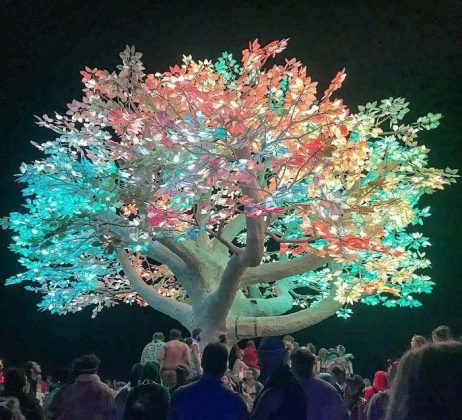 A large artificial tree with glowing leaves in vibrant pink, green, and blue stands against the night sky. Like a scene from Burning Man photos impossibles, crowds gather below, mesmerized by its brilliant lights and intricate branches.