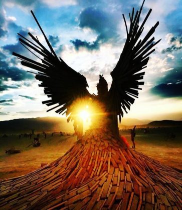 A dramatic wooden sculpture of a large winged figure stands outdoors at sunset, reminiscent of iconic BurningMan photos. The sun shines through the wings and scattered planks, while silhouetted people and bicycles dot the open landscape.