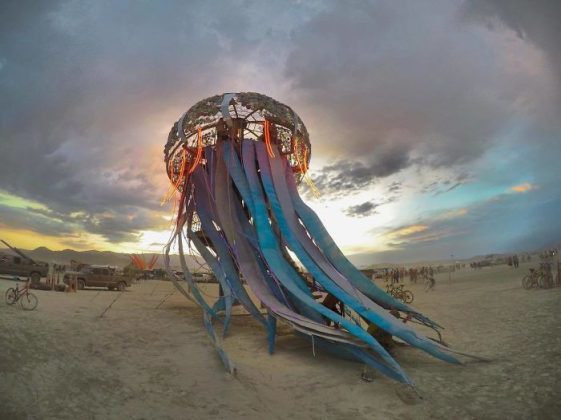 A large, artistic jellyfish sculpture with long, blue tentacles stands on a sandy desert at sunset. People and bicycles surround it as colorful lights glow—an iconic scene captured in BurningMan photos of surreal moments and photos impossibles.