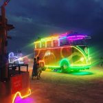 A brightly lit, neon-outlined vehicle resembling a Volkswagen bus drives across a sandy desert at dusk during Burning Man. The bus glows with rainbow lights, and festival-goers walk nearby. In the foreground is a “No Foot Traffic” sign edged in neon.