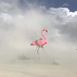 A giant pink flamingo sculpture stands on tall metal legs amid swirling dust, capturing the surreal essence of BurningMan photos. Two people sit on its back, partially obscured by haze, while cloudy skies shroud distant objects in the background.