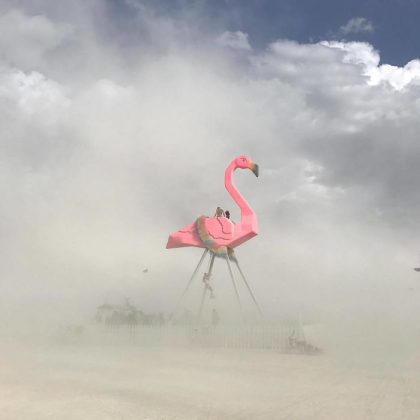 A giant pink flamingo sculpture stands on tall metal legs amid swirling dust, capturing the surreal essence of BurningMan photos. Two people sit on its back, partially obscured by haze, while cloudy skies shroud distant objects in the background.