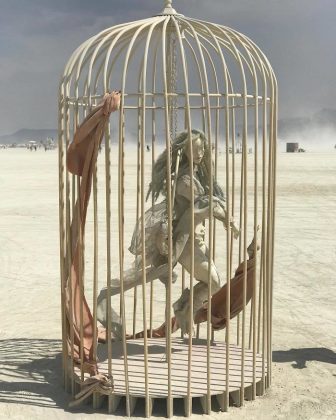 A life-sized, ghostly humanoid sculpture with long hair appears to run inside a large, pale birdcage at Burning Man. Torn brown fabric hangs from the bars. The cage rests on a barren, dusty plain under a hazy sky, with distant figures and mountains behind.