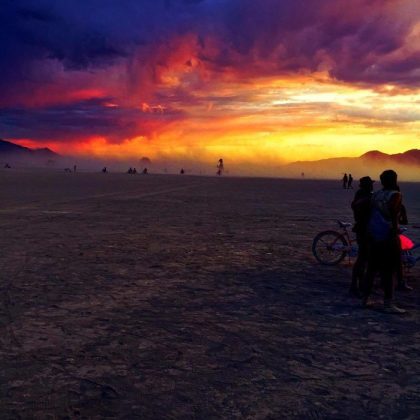 A dramatic sunset with vivid orange, yellow, and purple clouds lights up the vast desert at BurningMan. Silhouetted people and bicycles stand in the foreground, with others scattered beneath the colorful sky in these stunning BurningMan photos.