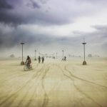 Cyclists ride across a vast, sandy desert under a cloudy, stormy sky at Burning Man. Tall, slender lampposts fade into the distance as dust and haze obscure distant art structures, creating photos impossibles in this surreal and ethereal atmosphere.