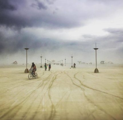 Cyclists ride across a vast, sandy desert under a cloudy, stormy sky at Burning Man. Tall, slender lampposts fade into the distance as dust and haze obscure distant art structures, creating photos impossibles in this surreal and ethereal atmosphere.
