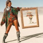 A woman in boots, vibrant top, scarf, and sunglasses stands on a desert playa at BurningMan, holding a large empty frame. Through it, a distant bike and art structure are visible—one of those unforgettable photos impossibles.