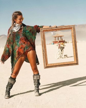 A woman in boots, vibrant top, scarf, and sunglasses stands on a desert playa at BurningMan, holding a large empty frame. Through it, a distant bike and art structure are visible—one of those unforgettable photos impossibles.
