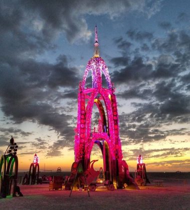A tall, ornate art installation arches upward, glowing with pink and purple lights against a dramatic sunset sky at BurningMan. The sculpture stands on a sandy expanse, surrounded by clouds and smaller lit structures—true photos impossibles.