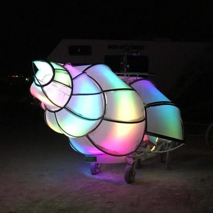 A large, illuminated sculpture shaped like a conch shell glows with soft gradients of rainbow-colored light. Reminiscent of iconic BurningMan photos, its metallic frame is visible beneath translucent panels, glowing vibrantly against the dark background.