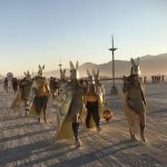 A group of people wearing white rabbit masks and gold capes walk together on a dusty, sunlit desert plain, evoking the surreal, festival-like energy seen in iconic Burning Man photos. Lamps, art installations, and distant mountains complete the scene.