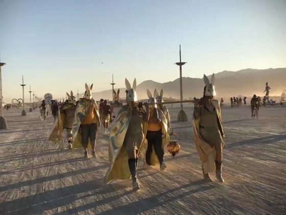 A group of people wearing white rabbit masks and gold capes walk together on a dusty, sunlit desert plain, evoking the surreal, festival-like energy seen in iconic Burning Man photos. Lamps, art installations, and distant mountains complete the scene.