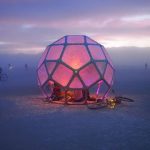 A glowing, geometric dome structure with translucent pink panels stands on a flat desert landscape at sunset—reminiscent of scenes from iconic Burning Man photos. Bikes are parked nearby and people relax beneath the dome as purple and orange skies stretch overhead.