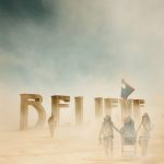 Three people in hats and goggles walk through a dusty desert at BurningMan, pushing a cart and holding a flag. Large sculpted letters spelling BELIEVE stand in the background, partially shrouded by dust under a blue sky.