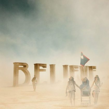 Three people in hats and goggles walk through a dusty desert at BurningMan, pushing a cart and holding a flag. Large sculpted letters spelling BELIEVE stand in the background, partially shrouded by dust under a blue sky.