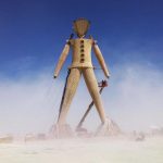 A giant wooden humanoid sculpture stands in a dusty, open desert beneath a clear blue sky. In classic Burning Man photos, workers on a cherry picker and scaffolding assemble or inspect the figure, secured by cables and braces at its base.