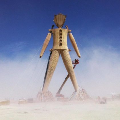 A giant wooden humanoid sculpture stands in a dusty, open desert beneath a clear blue sky. In classic Burning Man photos, workers on a cherry picker and scaffolding assemble or inspect the figure, secured by cables and braces at its base.