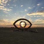 A large wooden sculpture resembling an eye stands on a flat, open desert at BurningMan. The sunrise or sunset shines through its center, illuminating a circular leafy wreath inside. Beams of sunlight radiate across the horizon—truly one for the photos impossibles.