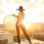 A person in a sheer, netted outfit and wide-brimmed hat stands on a rooftop at sunrise or sunset. Capturing iconic BurningMan photos, they hold out their garment as sunlight glows through it; vehicles and tents suggest a camp setting.