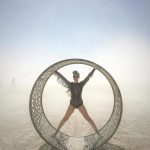 A woman in a black leotard and sunglasses stands with arms and legs outstretched inside a perforated metal ring on BurningMan’s dusty playa, creating one of those surreal, photos impossibles that capture the festival’s futuristic atmosphere.