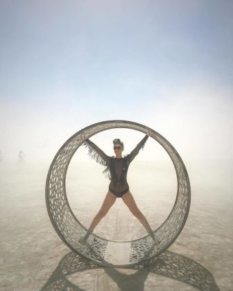 A woman in a black leotard and sunglasses stands with arms and legs outstretched inside a perforated metal ring on BurningMan’s dusty playa, creating one of those surreal, photos impossibles that capture the festival’s futuristic atmosphere.