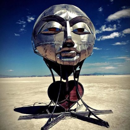 A large metallic sculpture of a stylized human face with calm features stands in a vast desert under a blue sky, recalling iconic BurningMan photos. The segmented head shines, supported by curved black metal, with distant mountains in the background.