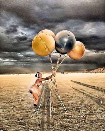 A person in festival attire and goggles stands on the sandy desert of Burning Man, holding large, colored balloons tied to poles. The cloudy sky is dramatic as balloon shadows stretch across the ground, with distant figures and structures beyond.