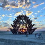 A large, intricate wooden sculpture stands on a desert playa at sunrise or sunset, glowing with sunlight through its center. Scattered clouds fill the colorful sky. Several bicycles and a few people evoke classic Burning Man photos on the sandy ground.