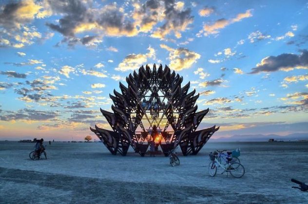 A large, intricate wooden sculpture stands on a desert playa at sunrise or sunset, glowing with sunlight through its center. Scattered clouds fill the colorful sky. Several bicycles and a few people evoke classic Burning Man photos on the sandy ground.