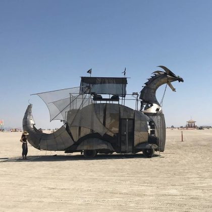 A large, metallic vehicle shaped like a dragon with an open mouth and spiked tail stands on a flat desert at BurningMan. Its body is made of panels and mesh, with a platform structure on its back. A person stands nearby for scale under the clear blue sky.