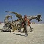 A person in silver, futuristic clothing stands before a massive metallic dragon sculpture made from scrap on a flat desert—an iconic scene among Burning Man photos, with distant people and tents under a clear blue sky.