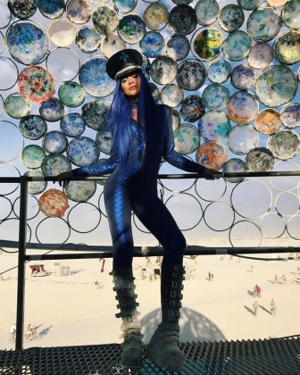 A person in a shiny blue bodysuit, tall platform boots, and a black hat with blue hair stands against a fence decorated with colorful plates at BurningMan. The background reveals a sandy landscape with scattered people and art installations.