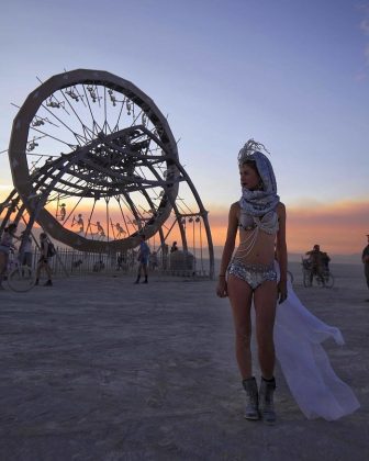 A woman in an ornate headpiece, bikini, and boots stands on a desert plain at sunset—a scene straight from BurningMan photos. Behind her, a massive metal wheel art structure teems with climbers as the sky glows orange and blue in these photos impossibles.