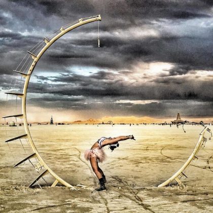 A person in festival attire bends backward gracefully under a large, crescent moon sculpture in the desert at Burning Man. The dramatic sky and distant art installations on the sandy playa create a scene worthy of photos impossibles.