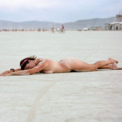 A woman lies face down on a vast, sandy landscape at BurningMan, her body dusted with sand. Wearing a pink flower headband and bracelets, she creates one of those photos impossibles against distant mountains and blurred structures under a cloudy sky.