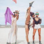Three women pose in a desert landscape at BurningMan. One lifts her leg high in a split, fringed outfit and boots flashing. The others, in sunglasses, smile—one in a sheer dress with hat and purple scarf, the other in mesh top and white shorts under cloudy skies.