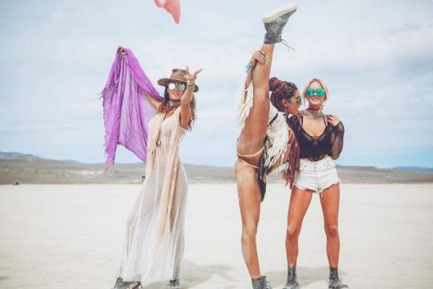 Three women pose in a desert landscape at BurningMan. One lifts her leg high in a split, fringed outfit and boots flashing. The others, in sunglasses, smile—one in a sheer dress with hat and purple scarf, the other in mesh top and white shorts under cloudy skies.