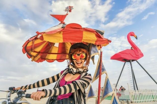 A smiling woman rides a bike with a colorful, striped umbrella hat at Burning Man. She wears large, flower-shaped sunglasses, a black-and-white scarf, and a pink top. In the background, there’s a giant pink flamingo sculpture and partly cloudy sky.