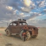 A woman in black boots and a bodysuit poses by a heavily modified, rusted Volkswagen Beetle with oversized off-road tires—an iconic BurningMan scene—parked on a desert plain under a dramatic, cloudy sky.