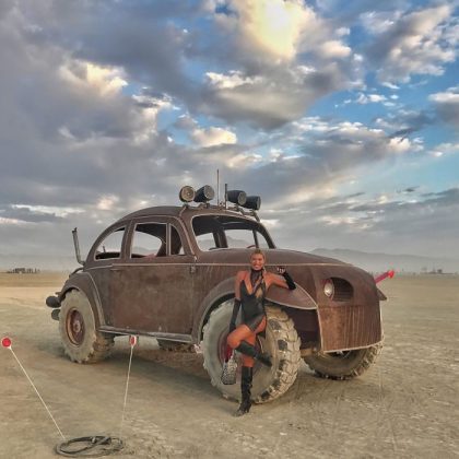 A woman in black boots and a bodysuit poses by a heavily modified, rusted Volkswagen Beetle with oversized off-road tires—an iconic BurningMan scene—parked on a desert plain under a dramatic, cloudy sky.