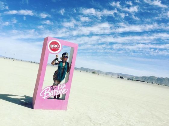A person stands inside a large pink box resembling a Barbie doll package with BRC Edition and Barbie written on it, channeling classic BurningMan vibes. Sunglasses and a hat complete their look in this iconic BurningMan photos scene under the vast desert sky.